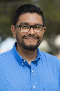 Picture of Joseph Duran, Administrative Assistant, Chicano Program. The image is a portrait of a man from the chest up. He is facing the camera and smiling slightly. The man has short, dark hair that's neatly combed back and sports a short beard and mustache. He is wearing dark-rimmed glasses and a bright blue button-up shirt with the top button undone. The background is blurred, indicating it might have been taken outdoors, with greenery and sky elements vaguely visible.